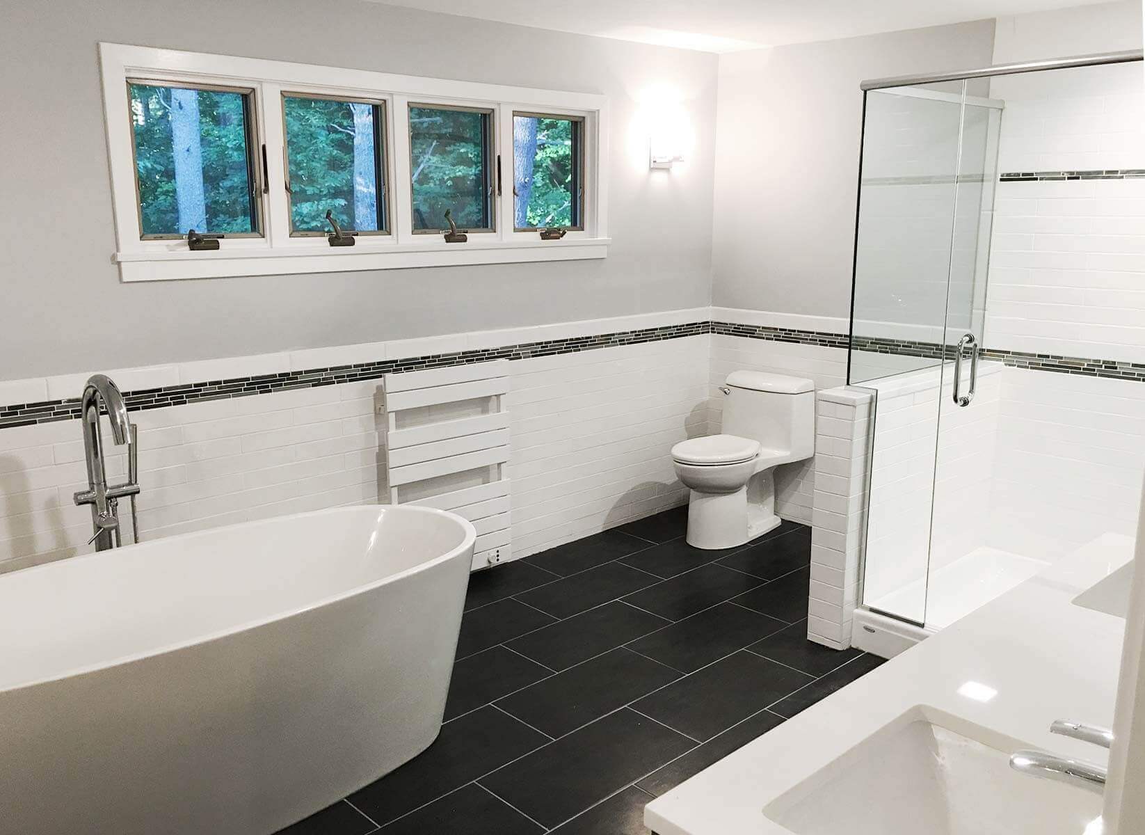 Large bath with glass shower, deep tub and sink in white with black tile floor.